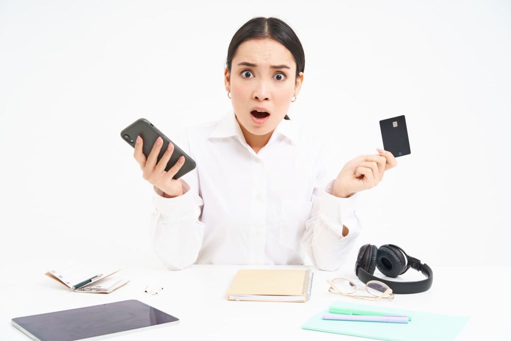 portrait-asian-woman-sits-table-holds-credit-card-smartphone-looks-upset-disappointed-by-p